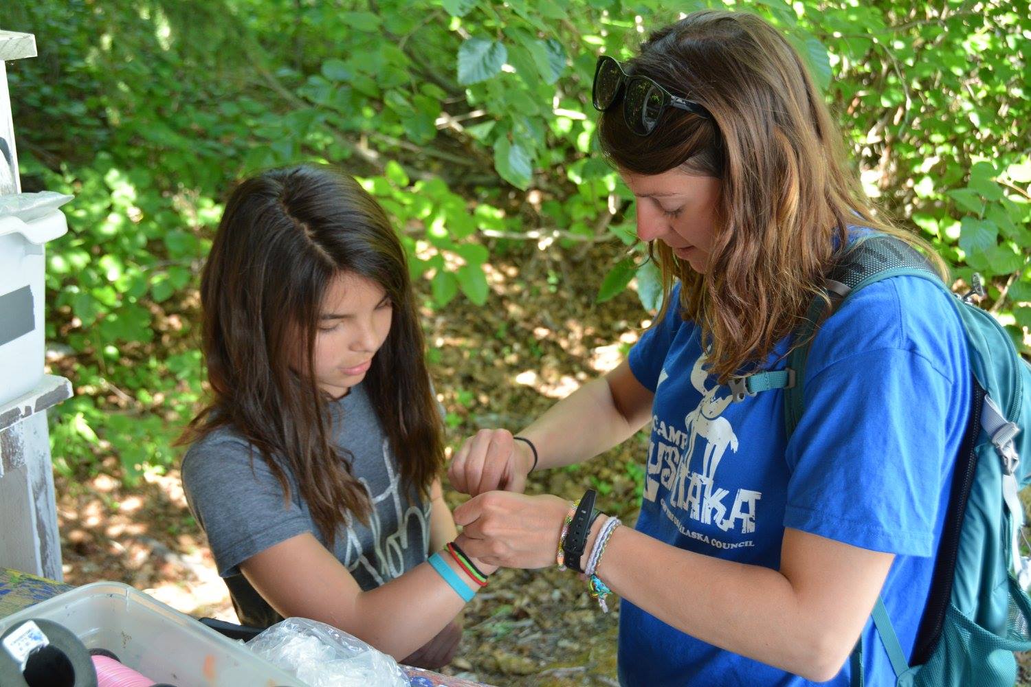 A young woman and a young girl, standing outside in front of green, leafy trees. The woman is helping the girl tie a bracelet on her wrist